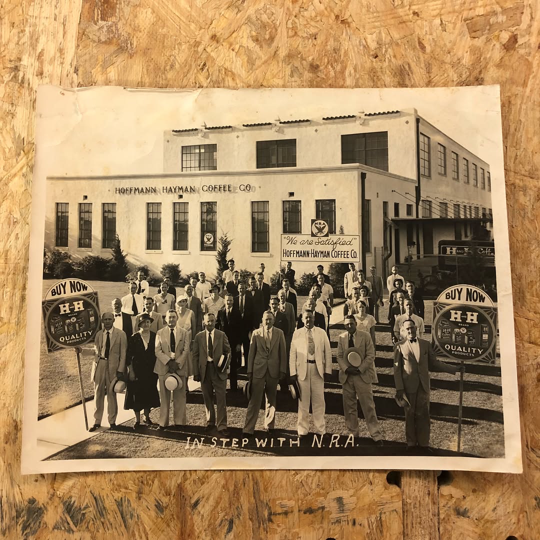 “In Step with N.R.A.” — Hoffmann-Hayman employees on the factory steps with Blue Eagle emblems, “We are Satisfied — Hoffmann-Hayman Coffee Co.” placard, and **IN STEP WITH N.R.A.** painted across the foreground; **circa 1933–1935** (collection press print).