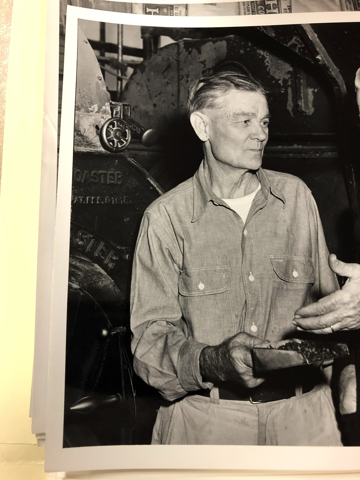 Black-and-white photographic print of a silver-haired middle-aged man in a light work shirt with rolled sleeves and a forearm tattoo, holding a sample pan of freshly roasted coffee beans in his left hand and gesturing toward a second figure just outside the frame, standing in front of a large patinated commercial drum coffee roaster with raised cast lettering reading "ROASTER" and "PAT." and a spoked cast hand-wheel on the feed door; the print is resting on a stack of other yellow-tone photographic prints.
