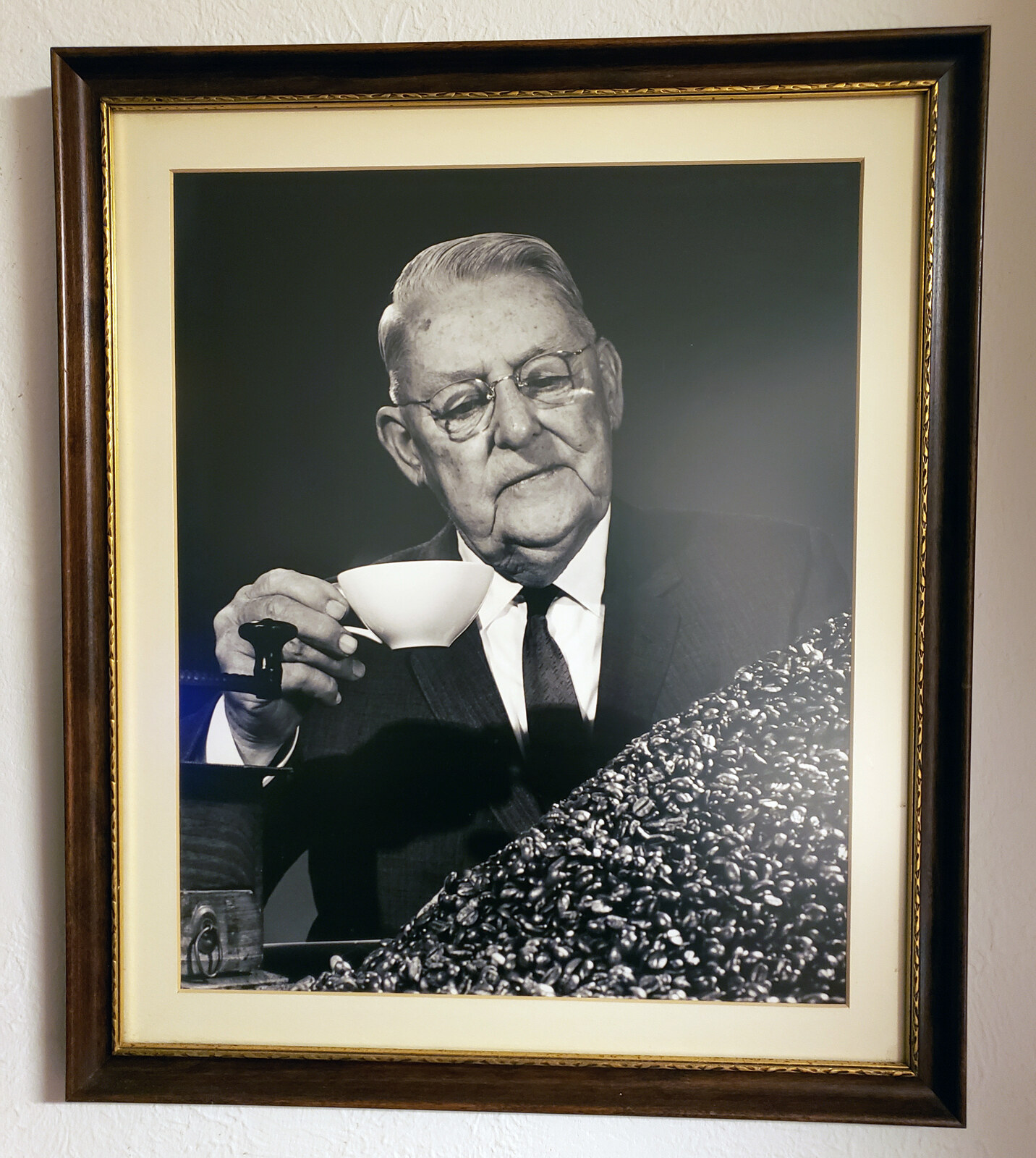Framed black-and-white studio portrait of an elderly heavy-set gentleman — wispy white hair combed back, round wire-rimmed spectacles low on his nose, white pocket square in a dark suit — holding a small white porcelain cupping cup near his face in his right hand and a black-handled cupping spoon in his left, looking down at a large cascading mound of whole roasted coffee beans on a polished dark bench with the cast-iron base of a shop-counter sample grinder at lower left; double-matted in cream in a dark-stained wood frame with gold-leaf trim, hanging on a cream-painted stucco wall. Subject tentatively identified as Gus P. Menger of Hoffmann-Hayman Coffee Co. in later life.