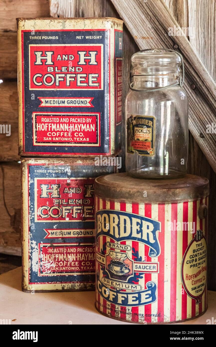 Color Alamy stock photograph showing an antique-shelf grouping against weathered wood-plank siding — two stacked three-pound rectangular H and H Blend Coffee tins at left in red, white, and blue with 'MEDIUM GROUND / ROASTED AND PACKED BY HOFFMANN-HAYMAN COFFEE CO. SAN ANTONIO TEXAS' labels in differing states of wear, a clear-glass wire-bail preserving jar with a 'WINNER Brand PICKLES - Packed by Geo. W. Wilson Co. San Antonio, Tex.' paper label standing in the center-top of the group, and at right a cylindrical red-white-and-blue vertical-striped Hoffmann-Hayman 'Border Brand Premium Coffee' pail whose ornate side cartouche plainly reads 'THIS BUCKET CONTAINS CUP & SAUCER PREMIUM / Hoffmann-Hayman Coffee Co. / San Antonio, Texas' — documenting the premium variant of Border Coffee called out on the brand page.