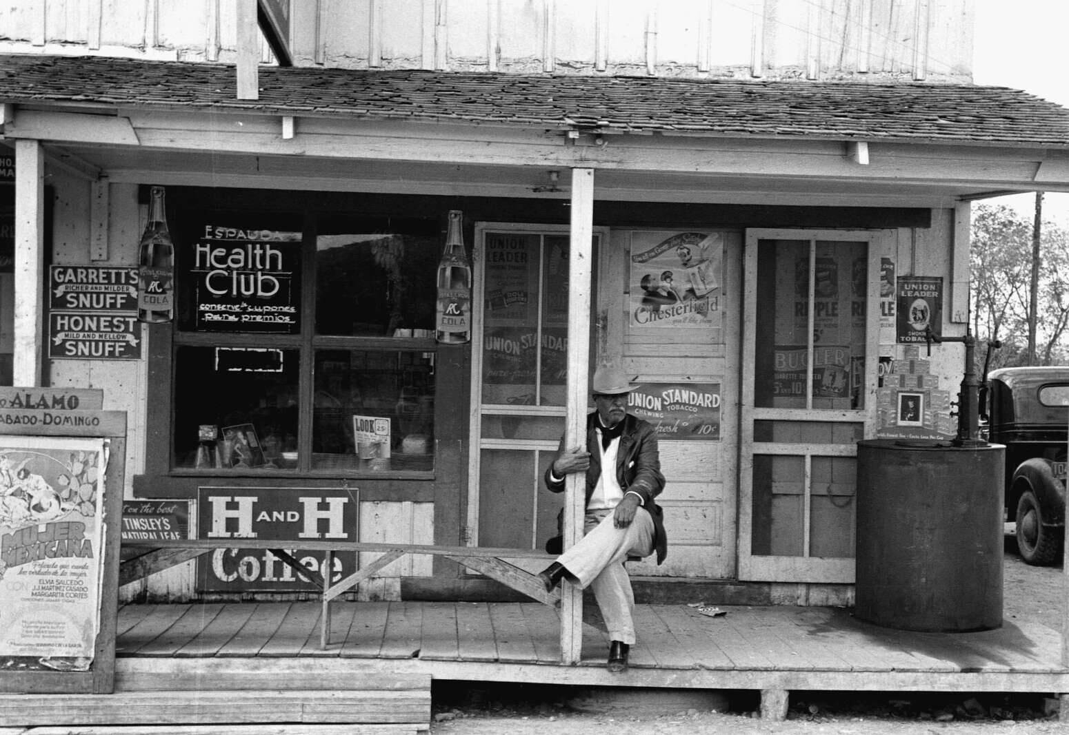 Black-and-white documentary photograph, in the manner of early-1940s Farm Security Administration / Office of War Information work, of a South Texas Tejano general-store porch — an older mustachioed gentleman in a pale fedora sits on the raised wooden porch against a storefront densely papered with period commercial signage: porcelain-enamel Garrett's and Honest snuff signs and tall RC Cola bottle-shaped advertising cutouts on the porch posts at left, a hand-lettered Spanish-language "ESPAUDA Health Club / Conserve su..." window sign and a "LOOK 25¢" magazine in the window display, through the door-glass Chesterfield and Union Leader and Union Standard and Bugler and Ripple tobacco ads, a Tinsley's Natural Leaf tobacco sign and a large dark-ground porcelain-enamel "H AND H Coffee" sign mounted on the low porch railing facing the street, and at the right a gravity-feed visible gasoline pump with the rear fender of a late-1930s automobile — and at the left edge a Spanish-language movie poster for the ALAMO theater reading MUJER MEXICANA / ELVA SALCEDO / J.J. MARTINEZ CASADO / MARGARITA CORTES, dating the frame to 1942 or shortly after.