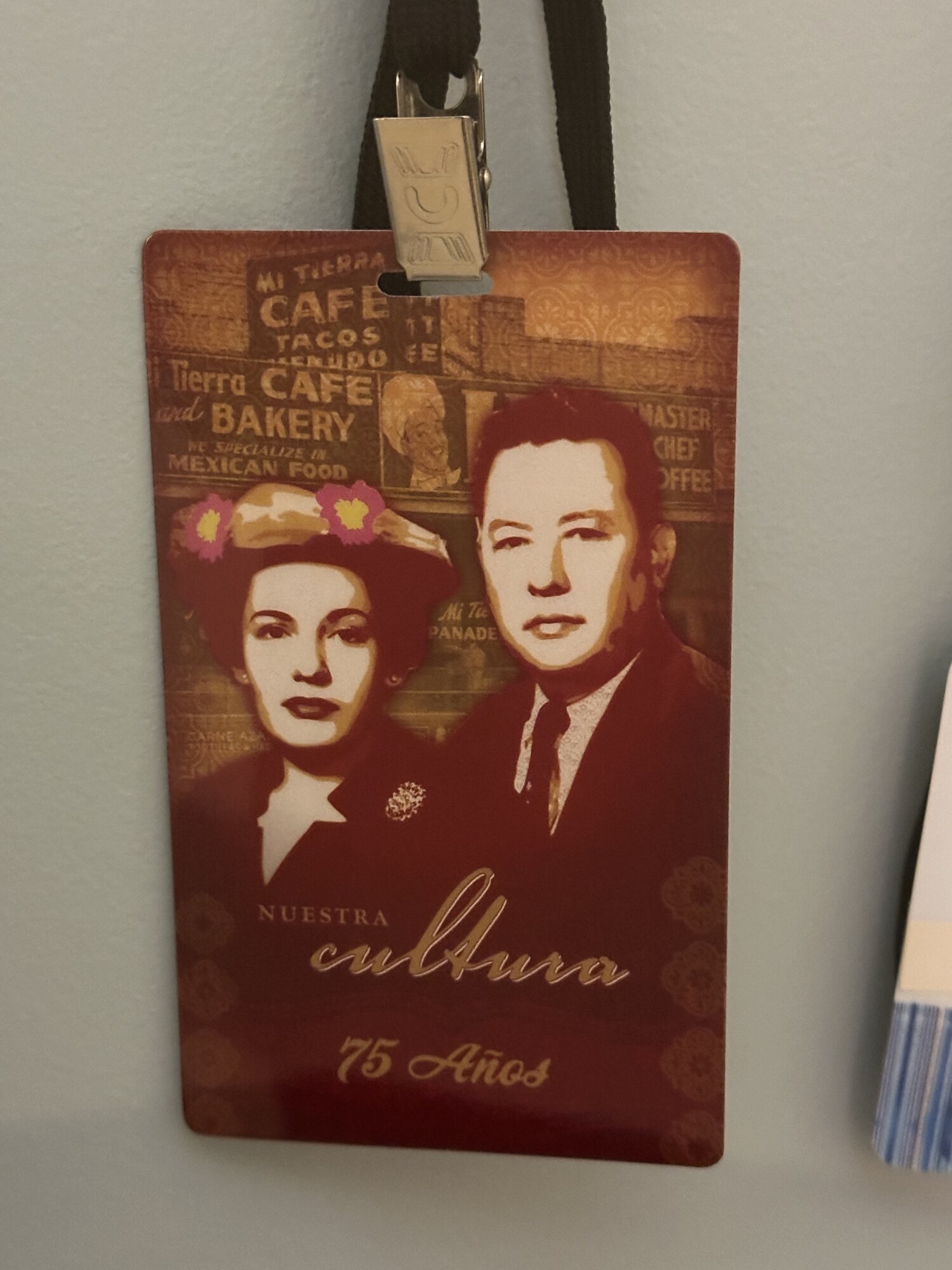 Color photograph documented 2024-01-23 at the Witte Museum, San Antonio, in the exhibit "Mi Cultura — Bringing Shadows Into the Light: The Photography of Al Rendón" — a vertical "Nuestra Cultura · 75 Años" lanyard pass from Mi Tierra Cafe & Bakery's September 2016 75th-anniversary celebration, almost certainly Al Rendón's own working press pass, hung from a black lanyard by a silver alligator-style metal clip on a pale exhibit wall, with the corner of an adjacent striped exhibit element just visible at right; the card carries sepia-and-burgundy stylized portraits of Pedro Cortez in a dark suit and Cruz Cortez in a hat with pink-and-yellow flower decorations against a montage of historical Mi Tierra storefront signage including (upper left) "MI TIERRA / CAFE / TACOS / MENUDO", (left of center) "Tierra CAFE / and BAKERY / WE SPECIALIZE IN / MEXICAN FOOD", (center) a small inset portrait of Doña Cruz Cortez, (center right) a small "Mi Tierra / EMPANADAS" reference, and (upper right) a vertical painted wall sign on a tile-and-stucco wall reading "MASTER / CHEF / COFFEE" — H and H Coffee's hotel-and-restaurant trade brand on Mi Tierra's storefront — with "Nuestra Cultura" in cream script and "75 Años" in cream below at the bottom of the card.