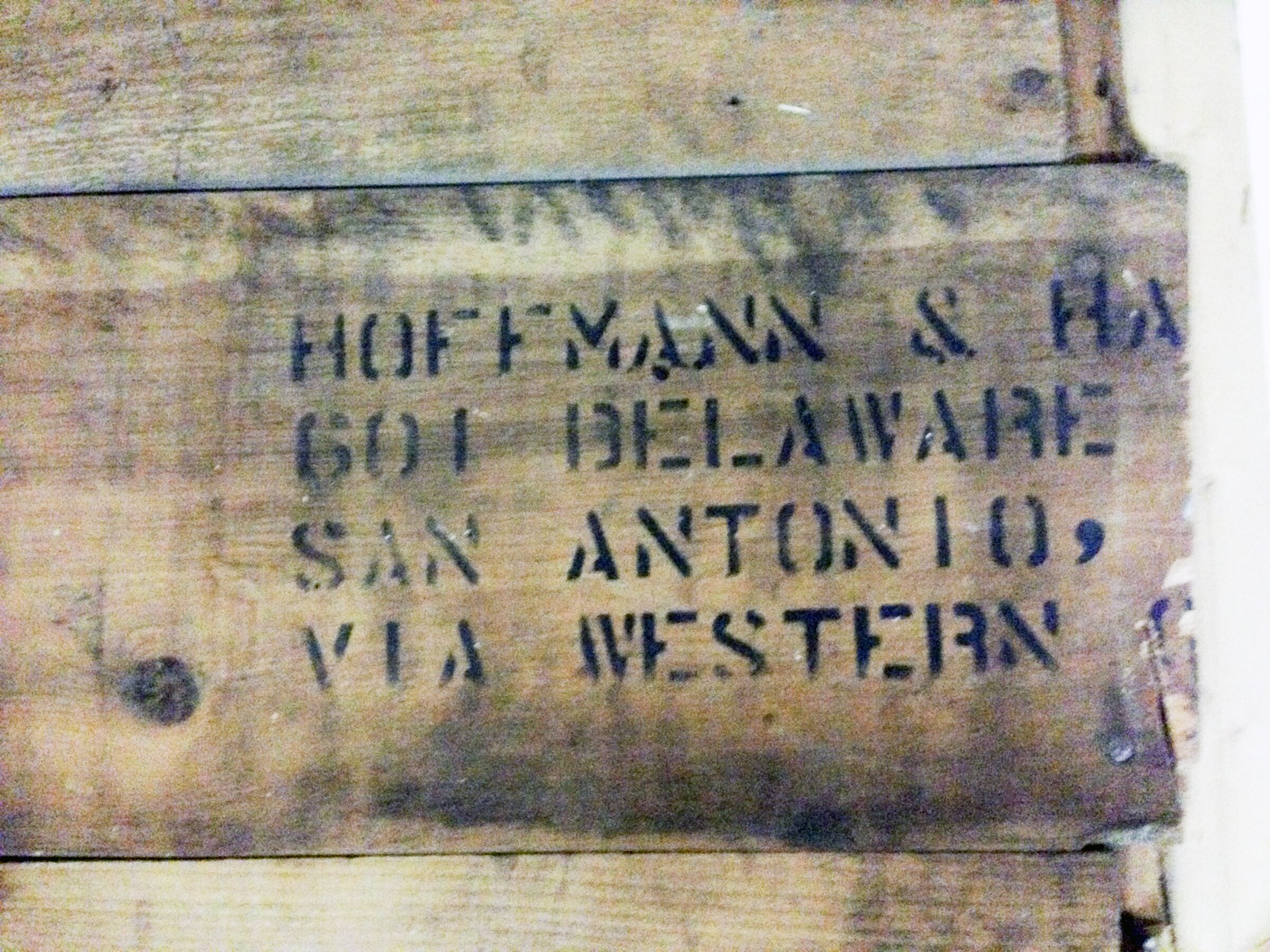 Horizontal crate board in a patchwork wood wall — black stencil reads HOFFMANN & HAYMAN (truncated), 601 DELAWARE, SAN ANTONIO, VIA WESTERN