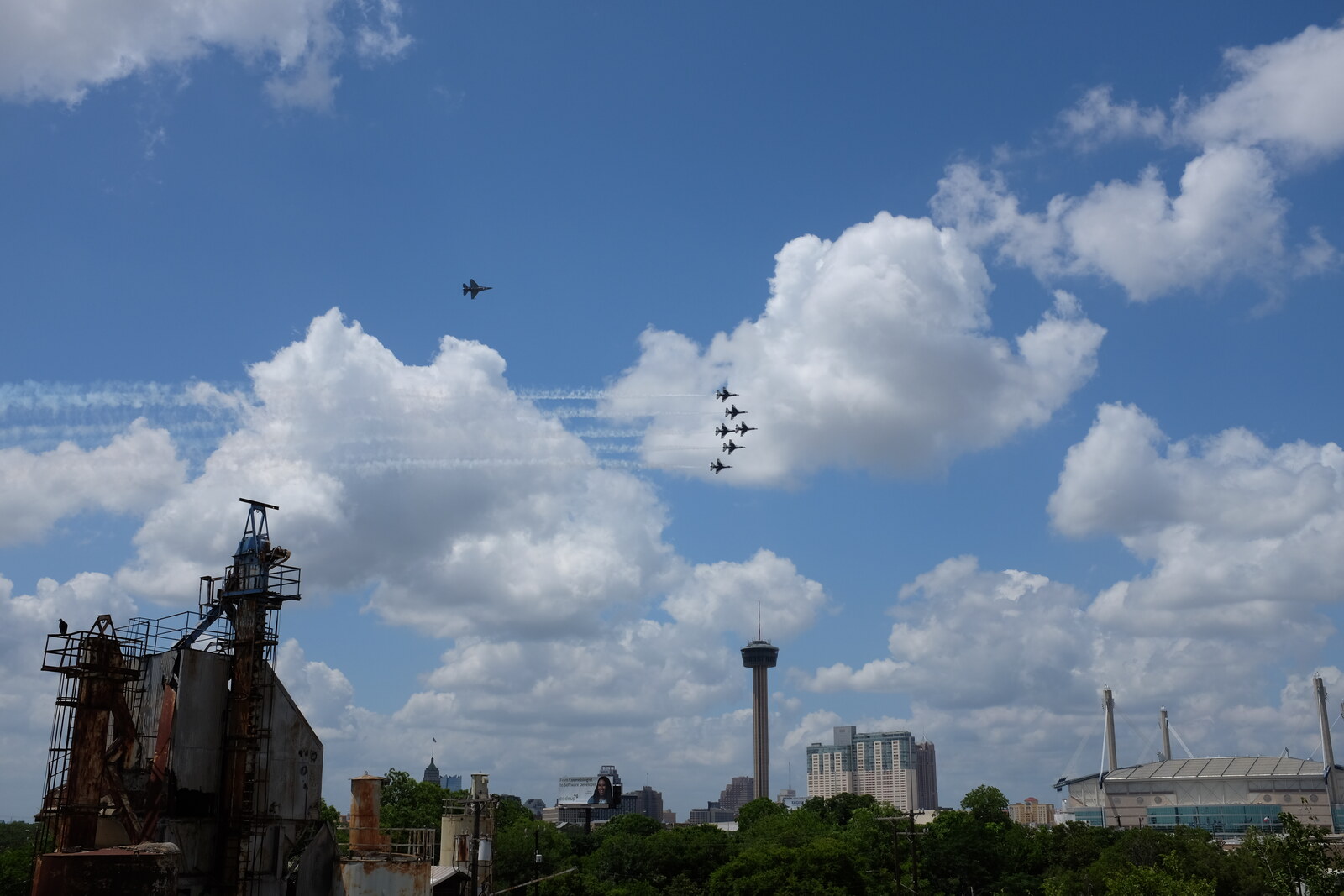 View north from the roof of the 1932 Hoffmann-Hayman Coffee Co. factory on 13 May 2020, with the plant's rust-brown coffee-silo headhouses in the lower-left foreground, the downtown San Antonio skyline and the Tower of the Americas at center, and the USAF Thunderbirds F-16 demonstration team in tight delta formation streaming contrails above the Tower with the solo ship running independently to the upper-left — the team's 'America Strong' COVID-era salute flyover.