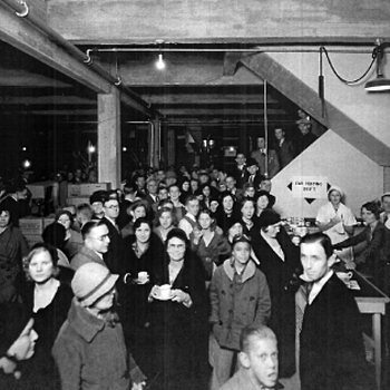 Black-and-white interior photograph, Hoffmann & Hayman Coffee Co. Delaware Street plant — dense crowd at the **December 1932** grand-opening week (GW Mitchell Construction archive image shared with the project). High industrial ceiling with beams; wall sign reading **CUP TESTING DEPT.** with arrow; guests in winter coats and hats hold small cups and saucers; white-uniformed staff at a serving counter at right. Reference documentation only — not an accessioned original negative in the museum files.