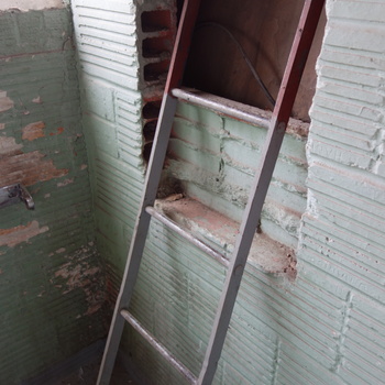 **22 February 2014** (**Sony DSC-RX100**) — upright metal ladder leaning through a **ragged breach** in a **pale green** painted **hollow structural tile** wall at the Hoffmann-Hayman factory, exposing **terracotta** cell faces and debris on the ledge; daylight from an upper opening.