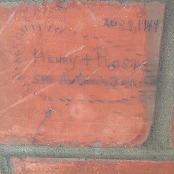Close-up, roughly top-down photograph of **one face of a reddish masonry brick** in **grey mortar** on an **interior wall of the second floor** at the Hoffmann-Hayman factory, documented **24 October 2014** (source **iPhone 5**) — the brick carries a **personal graffito** in dark ink or ballpoint: **'Henry + Rosa'** at center (with **'+'** joining the names), **'Aug 8, 1949'** filed in the upper right, and **'San Antonio, Texas'** centered below the names, all loosely enclosed inside a **dashed-line heart** outline drawn on the glazed brick face with a curved **arrow** running from lower-left toward the bottom of the heart; fine light **scratches** cross the brick face — a **six months later** doodle-on-the-wall **counterpoint** in English to the **February 1949 Spanish** tradesmen’s marks elsewhere in the same **upstairs expansion** chronology, but here reading as a **couple’s** hand rather than a crew date; joints to neighboring bricks visible at lower and right edges