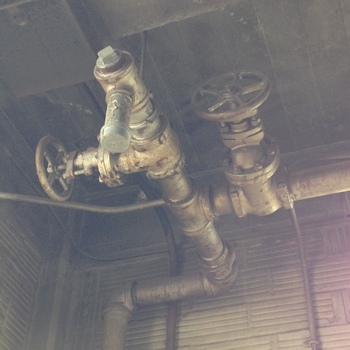 Upward-angled photograph on the **second floor** of the Hoffmann-Hayman factory — **gas piping** run **against the underside of the ceiling** (paired large **circular cast-iron gate-valve handwheels** bolted onto flanges on a horizontal galvanized or black iron header that Tee-branches upward to a capped vertical stub alongside elbows and screwed couplings); the metal carries decades of oxidation, flaky old paint residue, and plant grime typical of untouched upstairs mains; the upper field reads as rough dark plank or board ceiling soffit and the lateral wall backdrop shows corrugated or rib-pattern industrial cladding, with thin flexible cabling crossing behind mid-frame above a ribbed masonry or block-face lower wall — photographed **1 December 2014** (iPhone 5); **companions** elsewhere on second-floor plant services include the separately documented **[northwest-corner gas shutoff on a vertical riser](/assets/images/gallery/factory-upstairs-northwest-corner-gas-valve.jpg)** (5 January 2025), a physically different vantage on the upstairs gas installation