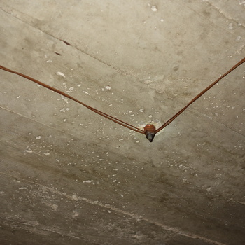 Looking up at the board-formed reinforced-concrete soffit in the space above the vault at the Hoffmann-Hayman factory, photographed 5 October 2014 (Sony DSC-RX100) — same low-clearance vault zone as the WATCH YOUR HEAD frame — horizontal wood-form grain, grey paste with pits and pale efflorescence speckling, and **two thin bare copper wires** entering from the upper left and upper right to meet in a shallow V at a **through-bolt or stud** with hex nut and washer countersunk into the concrete, a simple uninsulated branch or bonding jumper fixed directly to the cast ceiling