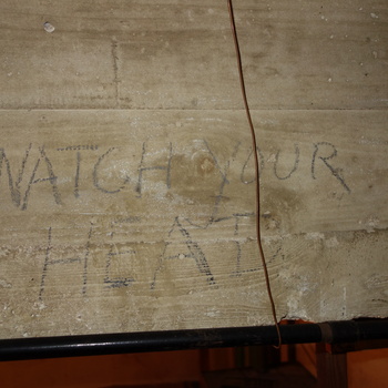 Low-clearance board-formed reinforced-concrete soffit or beam in the vault-adjacent circulation space at the Hoffmann-Hayman factory, photographed 5 October 2014 (Sony DSC-RX100) — horizontal wood-plank formwork grain pressed into the grey-beige concrete face, with the two-line hand lettering 'WATCH YOUR' / 'HEAD' applied in dark charcoal or grease stick; a thin copper wire hangs vertically through the field and a black horizontal pipe crosses the bottom edge, reading as a practical head-strike warning in tight quarters above the vault