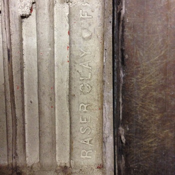 Close-up oblique photograph of structural clay tile or glazed hollow structural block in Hoffmann-Hayman factory masonry, **23 April 2015** (iPhone 5) — **light-grey** extruded ceramic face carved with parallel **deep vertical flutes** (narrow shadowed grooves alternating with pillow faces), **raised sans-serif stamping** spelling **FRASER CLAY CO.** (**CO.** abbreviated) reading **bottom-to-top** along one flute land; stray **blood-red aerosol/primer splatters** bead across ridges and grout lines; coarse **tan fibrous sheath** (**wood-fiber insulating board** or corrugated cover strip) kisses the unit at frame-right beside a pencil-thin mortar shadow gap — complements **[Fraser Clay terracotta block face photographed 24 October 2014](/assets/images/gallery/20141024-factory-fraser-clay-structural-tile.jpg)** in **this gallery** and ties to the hollow-tile masonry cross-section in **[the expansion hole](/assets/images/gallery/factory-upstairs-expansion-hole-through-original-exterior.jpg)**