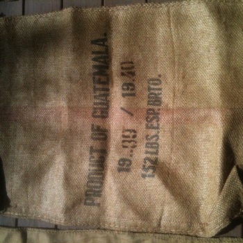 Burlap green-coffee sack on dark vertical wood planks — bold printed lines PRODUCT OF GUATEMALA; 1939 / 1940; 132 LBS ESP BRTO (weight and crop-era markings); faint red rule across sack — import sack form for Central American green coffee (not Hoffmann-Hayman lettered); project photograph 3 July 2015 for roaster sourcing context, another partial burlap sack at lower edge of frame