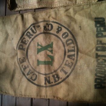 Burlap green-coffee sack on weathered wood planks — circular exporter stamp reading Cafe Peru and Co and N.B. Tealdo and Co around large green serif initials LX; bold PRODUCT OF PERU legend; smaller Lima Peru markings including Sacco Lazo Sudamericana — Peruvian import sack form typical of raw coffee shipped to U.S. roasters (not Hoffmann-Hayman printed); project photograph 3 July 2015 for sourcing context