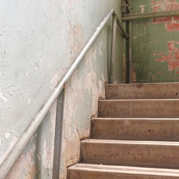 **1 August 2015** (**Fujifilm FinePix X100**) — service **concrete stair** with **galvanized pipe** handrail: peeling **mint-green** paint on plaster, **exposed red brick** on the landing wall, small pipes in the head — Hoffmann-Hayman circulation core.