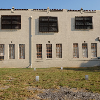 Bright **mid-morning** daylight photograph (**2 October 2015**, **Fujifilm X100T**) of the **full south elevation** of the Hoffmann-Hayman factory — textured **cream / buff stucco** over two stories under a flat roof with three **terraced parapet bays** capped in **Spanish / mission terracotta pantiles**, each ridge marked with a centred **small white finial**; **three** matching **steel-sash second-floor bays** screened by **welded diamond-mesh exterior security cages**, **nine** tall **segmented first-floor lights** (**three stacks of triple narrow bays**) behind **steel-bar grilles** with small rectangular **metal wall vents** above each stack; vertical **PVC downspout** between bays; foreground **thin grass**, **grey gravel**, and four **silver survey-style pins** punching a crude rectangle toward the façade; camera-right approaches the **Delaware Street entry** — concrete **steps**, pipe handrail, **flat-roof awning/canopy**, and a **white pedestal sign** carrying **601** in **red sans-serif numerals**, the published street-front number at the **Delaware-and-tracks gate** echoed in **[the 1932 *Sunday Light* Delaware plant clip]({{ site.baseurl }}/sunday-light-july-1932-factory-article-reprint/)**; further right clustered **meter socket / conduit penetration** spaghetti; extreme left beyond **chain-link** a wedge of **railroad-yellow rolling stock or maintenance vehicle** parallels the siding; **clear saturated blue sky** with stark **south-face sun shadows** tipping slightly **east** — complements **roof-edge context** **[southwest along the parapet, 1 Oct 2015](/assets/images/gallery/2015-10-01-factory-roof-south-parapet-looking-southwest.jpg)** and dusk ground views **[Feb 2019 trackside](/assets/images/gallery/factory-south-end-from-tracks-2019.jpg)** / **[Mar 2021 sunset](/assets/images/gallery/factory-south-face-sunset-2021.jpg)** on the **same south wall read**