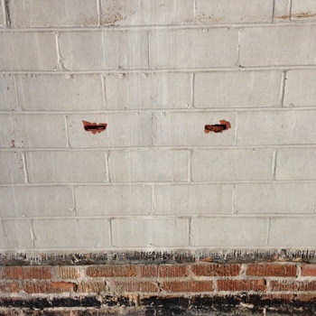 Second-floor **post-1932 expansion inventory** bay at the Hoffmann-Hayman factory — **west interior wall** of the **first expansion** shell (per site note: loading / storage zone), photographed **24 November 2015** (**iPhone 5**) — straight-on view of **painted concrete-masonry-unit (CMU) blockwork** with **two parallel horizontal ragged punctures** roughly **one block course high** and **two cells apart on center**, a damage pattern consistent with **lift-truck / forklift fork tines** having been driven through the wythe — distinct from the **deliberate oval exploratory hole** through the **original 1932 exterior leaf** documented **[in the adjacent frame](/assets/images/gallery/factory-upstairs-expansion-hole-through-original-exterior.jpg)**; the upper field shows **whitewash drip streaks** over grey block, a **rough mortar relieving band** below, and **exposed red perforated brick** with a **dark-stained** base course — **accidental equipment damage** to expansion-period interior masonry rather than a cut sample opening