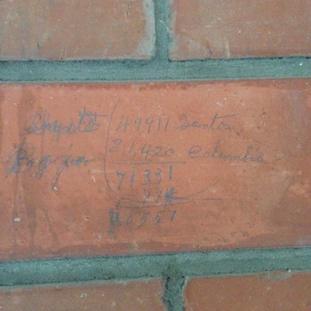 **Second-floor post-1932 expansion inventory** room — oblique photograph of **red clay brick wall** with **grey cement-lime mortar** joints (**24 November 2015**, **iPhone 5**), taken in the **same storage / loading bay** where **[fork-tip damage to CMU](/assets/images/gallery/2015-11-24-factory-second-floor-expansion-west-wall-forklift-fork-punctures.jpg)** is documented earlier in this gallery sequence — handwritten **coffee stock tally** scratched in **blue pencil** (with occasional strike-through corrections) straight on **brick bedding**: vertically stacked captions left of the tally read **‘Brazils / Bags.’** grouped lines show **Brazil Santos** and **Colombian** sack figures (with **comma-grouped totals** summed mid-field and a **revised figure** carried to the bracket foot), tying the architectural fabric to everyday **incoming green-coffee bookkeeping** beside the forklift-scarred masonry of the opposite wall.