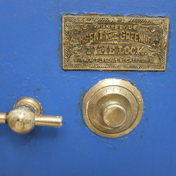**First floor**, **vault-zone circulation / plant office run** — close straight-on portrait of the **door face** of a **compact floor safe** stationed **outside the main vault enclosure** (**not inside** the large floor-safe chamber whose interior drawer faces are documented **[here](/assets/images/gallery/20150210-factory-vault-large-safe-drawer.jpg)** — distinct also from **[the wide-shot large safe casing](/assets/images/gallery/factory-large-safe.jpg)**), photographed **28 November 2015** (**Fujifilm X100T**) — **medium satin-blue enamel** door with **fine orange-peel** texture scuffed chip-white at flange edges; **rectangular ornate cast-brass manufacturer's tablet** centred high reading **'GUARDED BY THE'**, **'SARGENT AND GREENLEAF'**, **'TIME LOCK.'**, **'MANUFACTURED AT ROCHESTER, N.Y.'** (**Sargent & Greenleaf**) with darkened embossed recess fields; **brushed-metal combination index dial** with **0–100** graduation around the flange and **LEFT**/**RIGHT** inscription with curved directional arrows centred at twelve o'clock; **chrome-plated Tee handle** projecting left mid-field with oxidation pitting; overall reading as auxiliary **jeweller's / office strongbox** securing cash or documents beside heavier vault-rated equipment