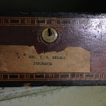 Close-up of one of the wooden interior drawers of the large floor safe inside the Hoffmann-Hayman factory vault, photographed 10 February 2015 (iPhone 5) — dark reddish finish with decorative gold-line banding, round brass keyhole at center-top, and an aged tan paper label typewritten with two lines reading 'MRS. C. B. MENGER' and 'INSURANCE'