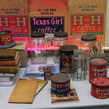 Overview of the Hoffmann-Hayman museum collection laid out on a white surface for display, photographed 21 May 2015 — rear row of large vintage paper H and H Coffee shipping sacks (12×2 lb and 24×1 lb regular) flanking a Texas Girl Coffee sack; midground cluster of clear glass jars (including Crystalvac-style handled jars), dark metal tins, and a thin bottle; foreground with stacked hardcovers, pamphlets, red Master Chef pound tins, blue-and-red H and H Coffee tins, and loose paper ephemera; a horizontal pink-violet accent light behind the glassware