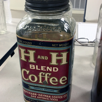 **iPhone photograph** **29 April 2017** — **clear** one-pound **H and H Blend Coffee** **Crystalvac** jar with **Crystalvac** script emboss on the shoulder, dark metal screw lid, and intact paper label reading **ONE POUND NET WEIGHT**, **H AND H BLEND Coffee**, **MEDIUM GROUND**, **HOFFMANN-HAYMAN COFFEE CO., SAN ANTONIO, TEXAS**, **STEEL CUT 100% PURE**, on a folding show table beside other glass — **Mac Johanson’s display** at the **Three Rivers Glass Show**, **Gurwitz Community Center**; same **Austin antique-shop** jar earlier documented in the **October 2016** forwarded Corpus Christi collector snapshot — **reference photograph**, physical jar **not** in the San Antonio museum collection