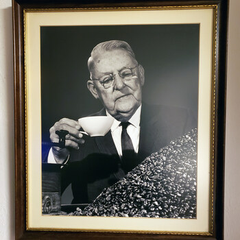 Framed formal studio portrait, black-and-white, of an elderly heavy-set gentleman with wispy white hair combed back, silver round wire-rimmed spectacles, a white pocket square, white dress shirt and patterned dark tie under a dark suit — holding a small white porcelain cupping cup near his face in his right hand and a black-handled cupping spoon in his left, looking down at a large cascading mound of whole roasted coffee beans on a polished dark bench with the cast-iron base of a shop-counter sample grinder (ring-pull catch drawer visible) at lower left, against a continuous studio backdrop — double-matted in cream with a gold inner accent in a dark-stained wood frame with gold-leaf trim, photographed hanging on a cream-painted stucco wall on 2019-12-04; subject tentatively identified as Gus P. Menger of Hoffmann-Hayman Coffee Co. in later life (identification not yet confirmed)