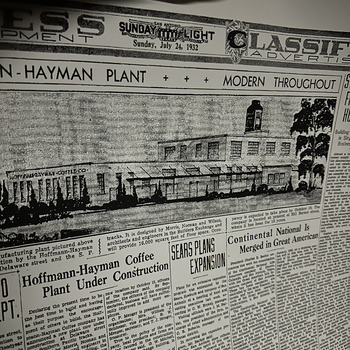 Large-format modern reproduction re-print of a page from the San Antonio Sunday Light, Sunday, July 24, 1932 classified / business-development section, photographed on 19 February 2023 lying flat on a light surface — across the top the paper's masthead reads 'SUNDAY LIGHT / Sunday, July 24, 1932' with 'CLASSIFIED ADVERTISING' on the right; immediately below, a banner headline spans the page reading 'HOFFMANN-HAYMAN PLANT · · · MODERN THROUGHOUT' and a large halftone architectural illustration of the new 1932 factory (captioned building signage 'HOFFMANN-HAYMAN COFFEE CO.' on the facade) shows the plant's full-length elevation with its central coffee-silo headhouse tower, the row of tall steel-sash industrial windows, and street-side trees — the building as originally designed before the second-floor expansion above the office was added; under the illustration runs the feature article 'Hoffmann-Hayman Coffee Plant Under Construction', which identifies the architects as Morris, Noonan and Wilson of the Western National (Builders Exchange) building, states the building will be fireproof reinforced-concrete construction with 16,000 square feet of floor space at Delaware Street and the Southern Pacific tracks, notes the company's then-current address as 331 Burnett Street (occupied for 10 years), lists the 1932 officers as G. P. Menger (president), Mrs. William J. Schlosser (vice president), R. W. Menger (secretary), and T. J. Menger (treasurer), and mentions both the planned rooftop 'Fragrant' advertising sign and the 'Crystalvac' coffee jar; adjacent unrelated articles headed 'SEARS PLANS EXPANSION' and 'Continental National Is Merged in Great American' are also visible on the page
