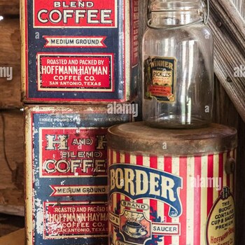 Color Alamy stock photograph (watermarked 'alamy') of a grouped antique-shelf display of San Antonio food-packing tins and a pickle jar against rough weathered wood-plank siding, documented 2023-02-25 — at left, two stacked three-pound rectangular H and H Blend Coffee tins in red, white, and blue (top label clean with only light wear, bottom one heavily paint-flaked and rusted) marked 'H AND H BLEND COFFEE / MEDIUM GROUND / ROASTED AND PACKED BY HOFFMANN-HAYMAN COFFEE CO. / SAN ANTONIO, TEXAS'; at right, a cylindrical red-white-and-blue vertical-striped 'Border Brand Premium Coffee' pail with a rusted press-fit lid, front cartouche showing a cup-and-saucer illustration with banners reading 'BORDER · BRAND · SAUCER · PREMIUM · COFFEE' and 'STEEL CUT' along the bottom edge, and a clearly readable ornate oval side cartouche reading 'THIS BUCKET CONTAINS CUP & SAUCER PREMIUM / Hoffmann-Hayman Coffee Co. / SAN ANTONIO, TEXAS' (establishing that this Border pail is an H and H product and a premium variant of the kind the Border Coffee brand page already calls out); and in the center of the group, standing on top of the Border pail, a tall clear-glass wire-bail preserving jar with a paper/foil label reading 'WINNER Brand PICKLES / SOUR PLAIN / CONTENTS 32 OZ. / Packed by Geo. W. Wilson Co. / San Antonio, TEX.' — not a Hoffmann-Hayman product but another early-20th-century San Antonio food-packing firm's jar, included here for contextual San Antonio packing reference