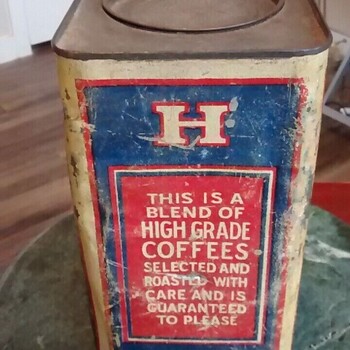 Color photograph documented 2023-05-20 of the same heavily worn large square-cross-section bulk-size H and H Blend Coffee tin photographed square-on to its opposite broad face — a deep blue field framed by a red double-line border on a cream ground, with a single cream-bordered red cartouche containing a serif 'H' centered above and a matching cartouche with a serif 'H' centered below, and at the center of the panel a large red rectangular cartouche with a cream double-line border carrying in cream serif capitals the full guarantee text reading 'THIS IS A / BLEND OF / HIGH GRADE / COFFEES / SELECTED AND / ROASTED WITH / CARE AND IS / GUARANTEED / TO PLEASE' — the same 'blend of High Grade coffees' product phrasing already documented as early-1920s H and H Blend language on the three-pound rectangular tin in the museum collection; surface heavily scuffed and scratched with paint loss along the bottom edge, the flat square top plate above with its circular recessed lid well visible (lid missing), set on a green marble side table against a wood-floor interior — not in the museum collection