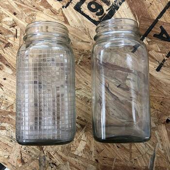 **Instagram** media export **17884975309229407** — **plan-view** photograph on a **tan OSB** workbench — **two empty crystal-clear square-section vacuum jars** (rounded-rect footprint, short **machine screw finish**, **lids absent**) presented **side by side** as **paired body variants** in the **Crystalvac** family Hoffmann-Hayman packed for **H and H Blend** and related lines: **left** jar carries a **waffle / raised-grid** mold texture wrapping the sidewalls from **shoulder** to **base**; **right** jar is **optically smooth glass** across the same profile; bright **specular highlights** skate across both shoulders; **upper-right** OSB shows **partial black stencil** characters (**6** or **9**, **A**) — **mold comparison** bracketing **[small Three Rivers Crystalvac pair](/assets/images/gallery/h-and-h-coffee-crystalvac-small-three-rivers-glass.jpg)** and the **[newspaper glass-show clipping](/assets/images/gallery/collection-newspaper-three-rivers-glass-show-h-h-blend-square-jars.jpg)** earlier in **this gallery**