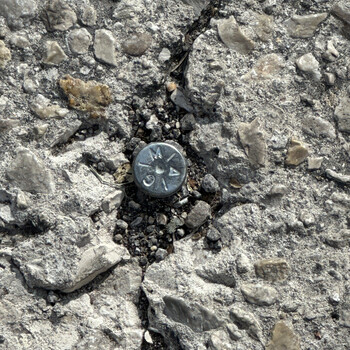 Close-up looking straight down at a small zinc/galvanized-steel surveyor's pavement marker driven into the asphalt-and-aggregate pavement of Delaware Street directly in front of the entry gate to the Hoffmann-Hayman factory, photographed on 23 December 2024 — a domed disk roughly the size of a US quarter sitting in a small crater of broken-out concrete and loose gray gravel, set into a pin grouted through the pavement, with a long vertical crack in the pavement running up through the marker's setting and continuing top to bottom through the surrounding aggregate; the face of the cap carries a stamped pattern of short radial tick marks emanating from a central center-punch dimple and three legible stamped letters arranged around the perimeter reading 'M' near the top, an 'A' (rendered as a triangular form, the crossbar faint) on the right, and a 'G' (a curled half-circle character) at the lower-left — most legibly read as 'G — M — A', a surveyor's initials, firm code, or agency stamp whose specific attribution is not yet identified; the marker sits on Delaware Street at the same Delaware-and-Southern-Pacific-tracks address recorded in the 1932 San Antonio Sunday Light feature on the new Hoffmann-Hayman plant ([documented in the Collection gallery](/sunday-light-july-1932-factory-article-reprint/)), a 21st-century property/survey artifact at the same street boundary the 1932 paper named when the factory was being built