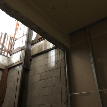 Standing at the north entry to the freight elevator on the first floor of the Hoffmann-Hayman factory and looking up through the board-formed concrete shaft header into the second floor above — concrete block (CMU) shaft walls, vertical steel elevator guide rails, and electrical conduit run up to the opening, with wood framing and scaffolding visible through the bright daylight at the top