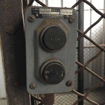 Tight close-up of the UP/DOWN pushbutton station controlling the **smaller second lift** at the Hoffmann-Hayman factory — a separate, smaller mechanism from the building's main interior freight elevator, fitted with an **exterior access door opening to the track side** so loads can be moved directly between the plant and the Southern Pacific siding running along the west flank — a grey-painted cast-iron pushbutton enclosure made by **Square D Company, Milwaukee, Wis., U.S.A.**, brass nameplate stamped 'CLASS 9001 / TYPE B-10 / MAX. V. 600 AC/DC / 218278 GG', two heavy round push buttons each cast with the legends 'UP' (top) and 'DOWN' (bottom), the button faces worn dark and oily from decades of use, mounted by four corner machine screws to a riveted steel angle-iron post behind a heavy diamond chain-link guard mesh — same physical Square D station documented in the wider track-side-lift cage view (matching nameplate and serial 218278 GG)