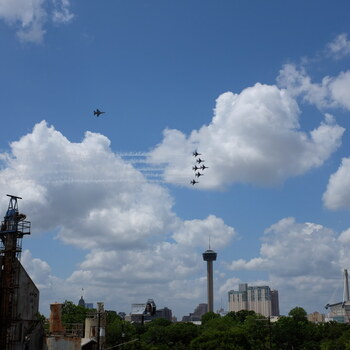 View from the Hoffmann-Hayman factory roof on 13 May 2020, looking north across San Antonio as the USAF Thunderbirds perform their 'America Strong' COVID-era salute flyover — the rust-brown tops of the factory coffee silos and a catwalked headhouse fill the lower-left foreground, green tree canopy stretches across the middle distance, and the downtown skyline rises through the trees with the Tower of the Americas at center, the Alamodome's tiered fabric roof at far right, and a civic dome peeking above the trees on the left; six F-16s stream contrails in a tight delta formation above and just right of the Tower of the Americas, with the Thunderbirds solo ship running independently higher and to the upper-left, all set against a brilliant blue partly-cloudy sky
