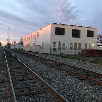Dusk exterior of the 1932 Hoffmann-Hayman Coffee Co. factory in San Antonio, photographed in February 2019 from between the parallel rails of the adjacent siding looking north — the south end wall of the two-story white-stucco plant catches the pink sunset on its terracotta-pantile parapet coping and corner finials, and the full length of the west (rail-side) flank recedes into shadow showing the row of tall multi-pane steel-sash industrial windows on the second floor above smaller barred first-floor openings, chain-link fencing, agave plantings, and a red dumpster along the yard