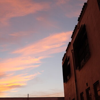 Dusk exterior of the Hoffmann-Hayman Coffee Co. factory's south face at 8:16 PM on 14 March 2021, photographed from the south looking north — the south end wall of the two-story cream-stucco plant recedes to the right of the frame in warm ambient shadow (near the spring equinox the sun sets close to due west, so the south face receives only ambient glow at dusk), with the terracotta pantile parapet coping stepping across the top edge, caged second-floor multi-pane steel-sash industrial windows and first-floor openings both covered with bolted-on diamond-mesh security grilles and glowing from within with warm yellow interior light, and a vertical downspout running between the upstairs and downstairs window bays — set against a dramatic pink, coral, and orange sunset sky with wispy cirrus streaks filling the open west side of the frame, a small silhouette element just visible on the horizon at bottom centre