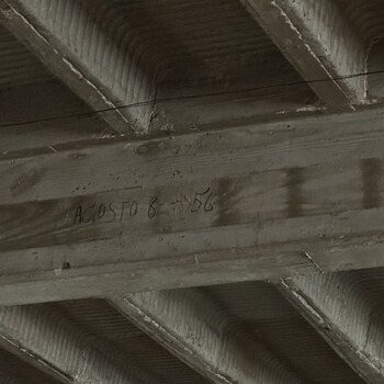 Upward view of the ceiling soffit in the second-floor expansion room above the office at the Hoffmann-Hayman factory, photographed on 1 February 2023 — the building's 1930s pan-formed reinforced-concrete joist floor system shown left honestly exposed with no suspended ceiling: flat-soffit concrete joists (ribs) run in parallel across the room with board-formwork grain pressed into their vertical sides from the wood-plank forms they were cast against, and the bays between the joists carry the characteristic ribbed soffit pattern left by removable corrugated-steel pan forms that were set between the joist forms, poured over, and stripped out after curing; on the flat face of the nearest joist a cursive graphite-pencil inscription reads 'AGOSTO 8 - 56' (August 8, 1956), a fourth dated pencil mark by the same hand or cohort as the three 'Marzo' 1952–54 dates on the east wall of the same room, and a thin black signal cable runs horizontally across just below the joist