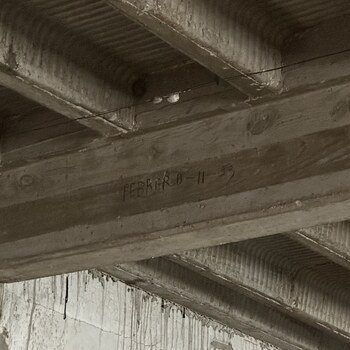 Upward view showing the junction between the pan-formed reinforced-concrete joist ceiling of the second-floor expansion room above the office at the Hoffmann-Hayman factory and what was originally a 1932 exterior masonry face of the plant (now an interior wall enclosed by the expansion), photographed on 1 February 2023 — in the upper two-thirds, parallel flat-sided concrete joists run across the frame with board-formwork grain and round knot-hole imprints pressed into their sides, and the bays between them carry the characteristic ribbed soffit pattern left by removable corrugated-steel pan forms; on the flat face of one central joist a cursive graphite-pencil inscription reads 'FEBRERO - 11 - 53' (February 11, 1953), a second pencil date by the same hand or cohort as the three 'Marzo' 1952–54 dates on the east wall of the room and the later 'Agosto 8 – 56' mark on the adjacent ceiling joist; below the ceiling, the top of the former exterior masonry face of the original 1932 plant meets the expansion soffit, its painted surface streaked with long vertical whitewash drip-runs from when the new interior ceiling above was painted, and a wooden jamb is visible at lower-right in what appears to be a post-expansion cut opening in the old exterior wall; a thin black signal cable runs horizontally below the inscribed joist