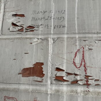 Close-up of the east interior wall of the second-floor expansion room above the office at the Hoffmann-Hayman factory, photographed on 1 February 2023 — scored plaster/render finished in white paint that imitates brick coursing (raised mortar-joint lines running horizontal and vertical across the surface), the paint delaminating and peeling extensively in irregular sheets and a large T-shaped spall at center-left revealing the warm red-brown substrate underneath; three cursive graphite-pencil inscriptions centered in the upper field, written in the same hand in Spanish — 'Marzo 5 - 1952', 'Marzo 25 - 1953', and 'Marzo 18 - 1954' — three mid-March dates one per year across three consecutive years, someone returning to this wall each March; additional red crayon / lumber-keel markings in the lower half including large peaked/triangular shapes on the right and partial red letterforms (an 'R' and the start of another character) at the bottom edge