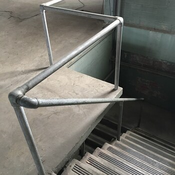 Top of the stair leading down from the second floor at the Hoffmann-Hayman factory, guarded by a galvanized-steel pipe handrail assembled from threaded elbow and tee fittings; ribbed cast-iron treads descend through the concrete floor slab, with a teal-painted wall and exposed brick visible behind
