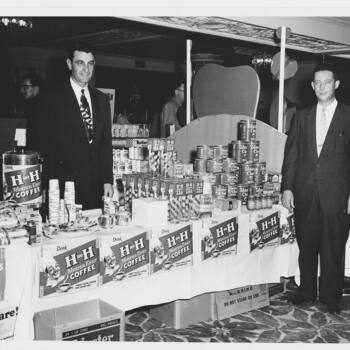 **Black-and-white photograph** from **Instagram** media export **17862082333188116** — Hoffmann-Hayman **exhibit or hotel-ballroom sampling table** (**not** a photograph of an object **in the museum collection**) — two **men in dark mid-century suits and ties** stand behind a **long white-skirted** counter in an **ornate public room** (patterned carpet, **crystal chandelier** and **ceiling mouldings** soft-focused behind); **foreground left**, polished **metal coffee urn** lettered **'Drink H-H Master Chef Coffee'** beside **stacks of paper cups** and **sample plates**; **skirted fascia** carries repeated **chef-head portrait** cards reading **'Drink H-H Master Chef Coffee — Finest Hotel Coffee For Home Use!'**; **center**, tall piles of **H-H Tea** folding cartons; **right**, **pyramided retail stacks** of **Master Chef Coffee** litho **one-pound** cans; under the cloth, **corrugated shipping cases** including one marked **'24 - 1 LB. CANS'**, **'VAC. PACKED'**, **'WARNING DO NOT STAND ON END'**; **stylized scenic backdrop** board behind the product mass — documents **co-marketing of Master Chef Coffee with H and H Tea** in a **trade-show / hotel catering** posture